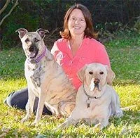 Debi McKee sitting outside between her two dogs, Bear (a yellow lab) and Ginger (a mixed breed dog).