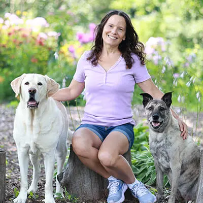 debi-mckee-ginger-bear-2025x200 Debi McKee sitting outside between her two dogs, Bear (a yellow lab) and Ginger (a mixed breed dog).